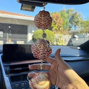 May include: A hanging car ornament featuring three vertically aligned scallop shells with reddish-brown patterns. The shells are connected by thin gold-toned metal chains, small white beads, and a circular metallic charm between the top two shells.