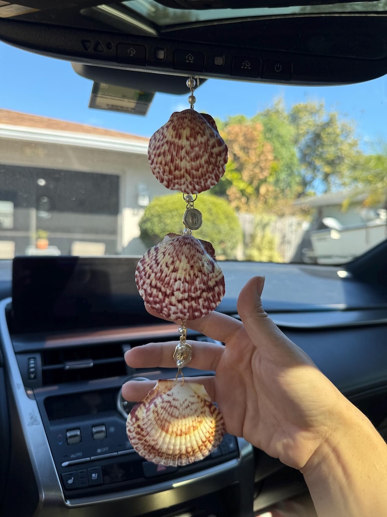 May include: A hanging car ornament featuring three vertically aligned scallop shells with reddish-brown patterns. The shells are connected by thin gold-toned metal chains, small white beads, and a circular metallic charm between the top two shells.
