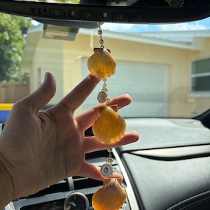 May include: A car rearview mirror hanging ornament featuring three orange scallop shells connected by a gold-toned chain with small white faux pearls and two small circular charms with white enamel and gold-toned star accents.