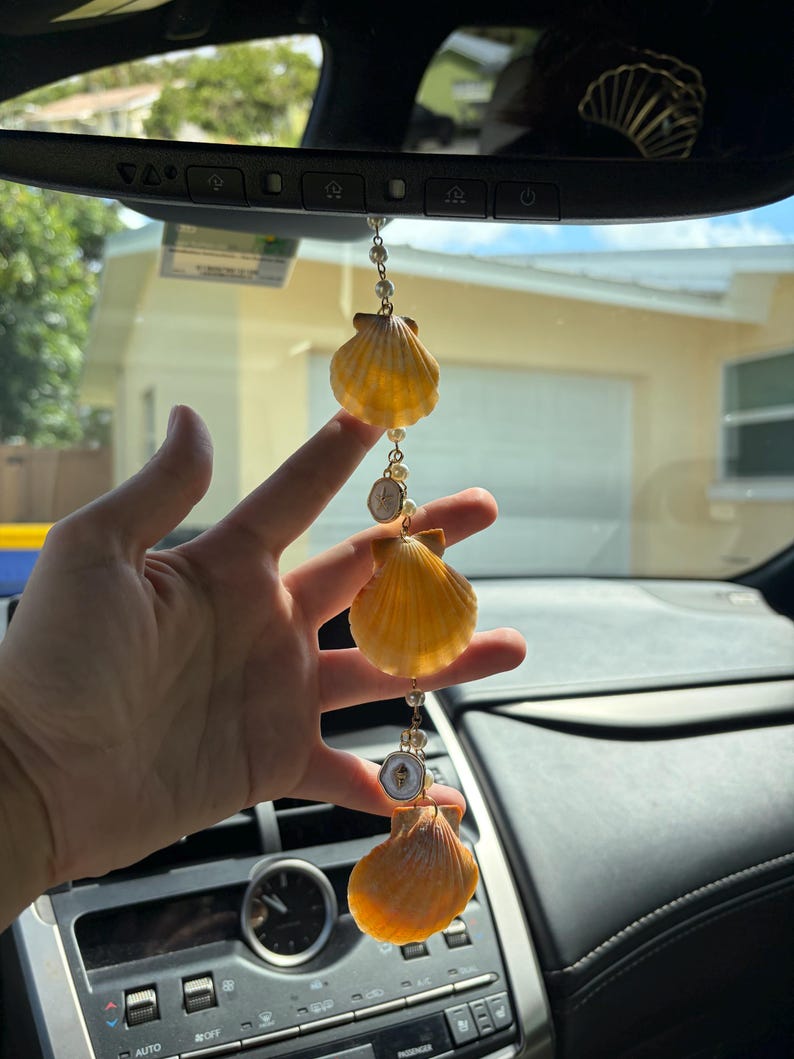 May include: A car rearview mirror hanging ornament featuring three orange scallop shells connected by a gold-toned chain with small white faux pearls and two small circular charms with white enamel and gold-toned star accents.
