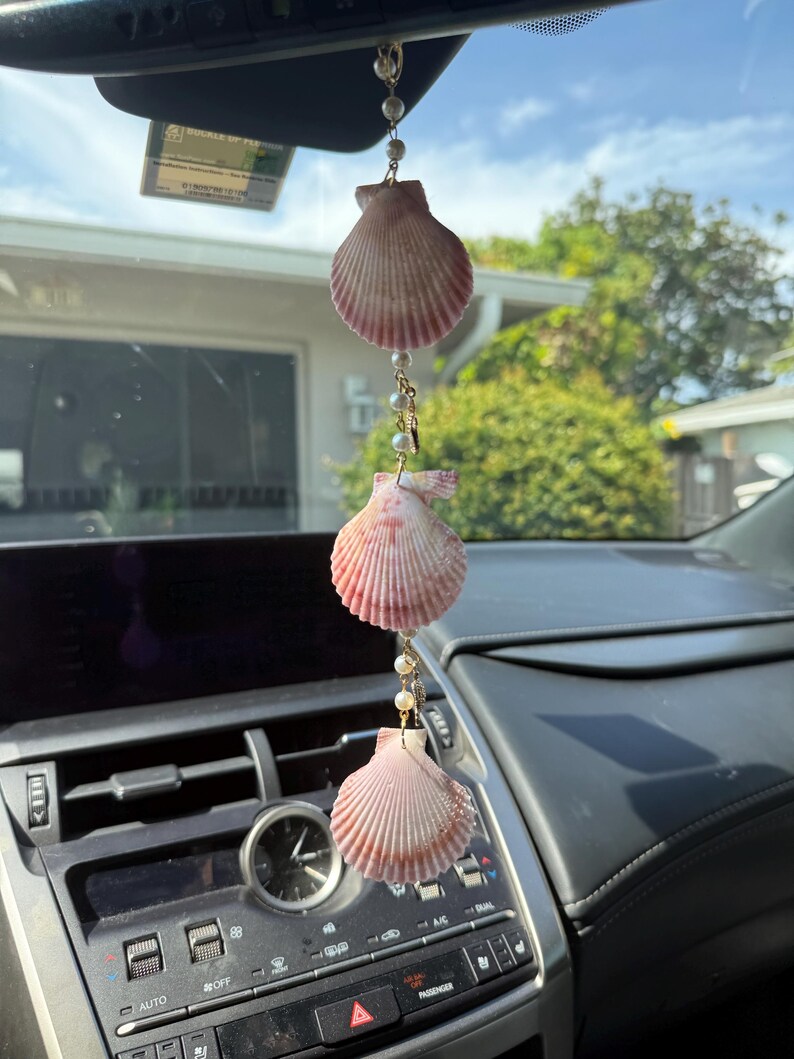 May include: A hanging car ornament featuring three pink and white scallop shells arranged vertically, connected by gold-toned metal links and small white beads. The decorative piece is suspended from a rearview mirror by a thin chain.
