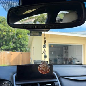 May include: A car rearview mirror hanging ornament featuring a vertical gold-toned chain with small circular accents, a starfish charm, and a large, textured, fan-shaped seashell at the bottom, suspended by a thin metal link chain.