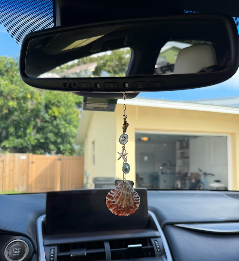 May include: A car rearview mirror hanging ornament featuring a vertical gold-toned chain with small circular accents, a starfish charm, and a large, textured, fan-shaped seashell at the bottom, suspended by a thin metal link chain.