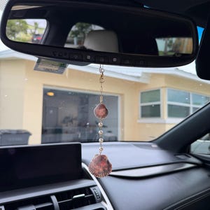 May include: A car rearview mirror hanging ornament featuring a gold-toned chain, a small brown seashell at the top, a series of white beads in the center, and a larger brown seashell at the bottom, suspended from the mirror.