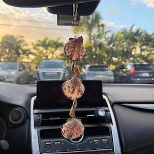 May include: A hanging car ornament featuring three fan-shaped seashells with reddish-brown patterns, interspersed with smaller tan-colored shells. The shells are connected by a thin gold-toned metal chain, designed to hang from a rearview mirror.