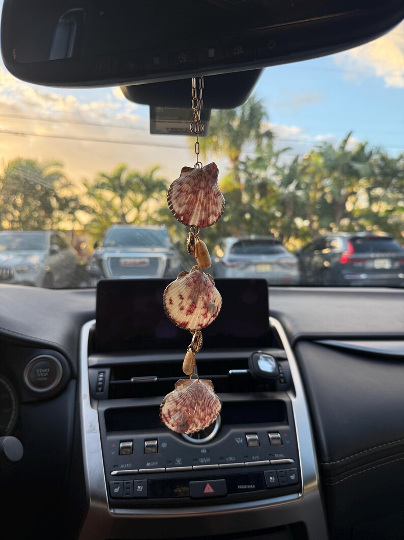 May include: A hanging car ornament featuring three fan-shaped seashells with reddish-brown patterns, interspersed with smaller tan-colored shells. The shells are connected by a thin gold-toned metal chain, designed to hang from a rearview mirror.