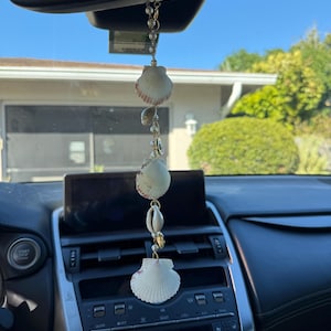 May include: A hanging car ornament featuring a vertical chain of white seashells, small white pearls, and gold-toned metal accents. The decorative piece is suspended from a rearview mirror, showcasing three distinct fan-shaped shells and a small cowrie shell.
