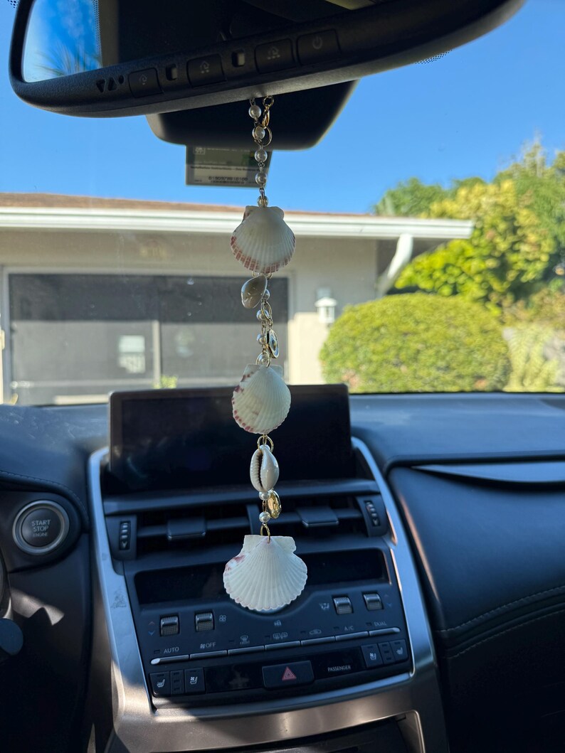 May include: A hanging car ornament featuring a vertical chain of white seashells, small white pearls, and gold-toned metal accents. The decorative piece is suspended from a rearview mirror, showcasing three distinct fan-shaped shells and a small cowrie shell.