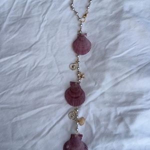 May include: A vertical hanging ornament featuring three purple scallop shells interspersed with white faux pearl beads on a gold-toned chain. The design includes small gold-toned charms, including a starfish, a heart-embossed disc, and a shell.