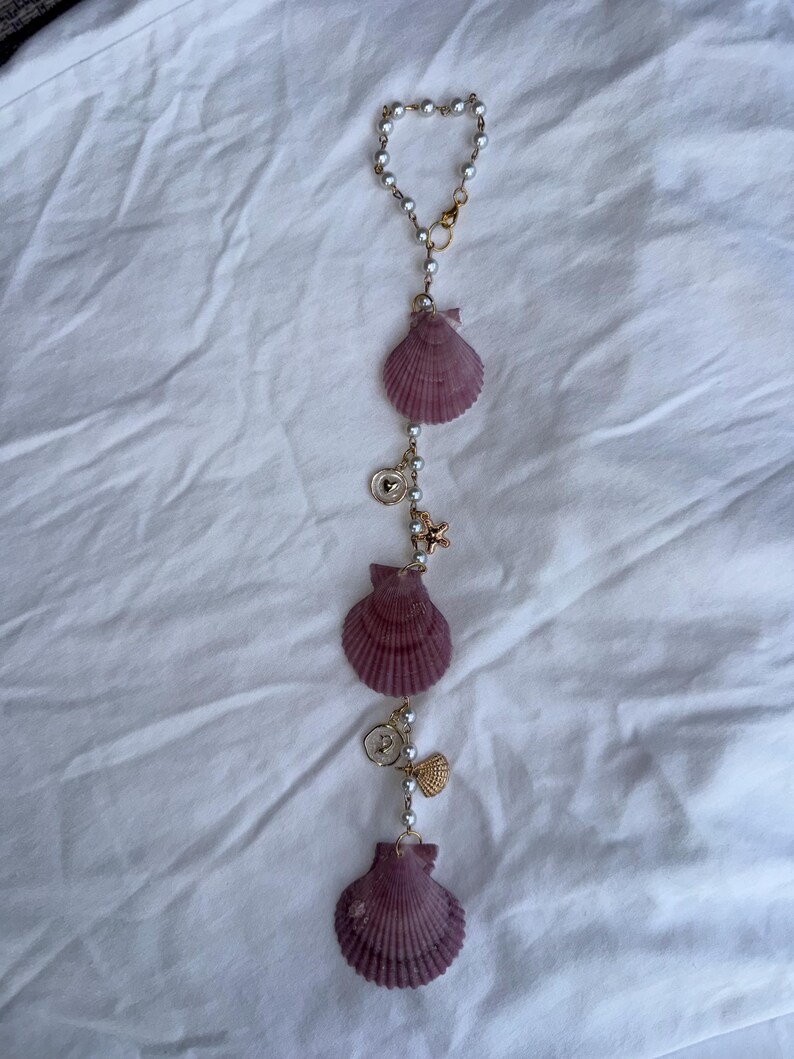 May include: A vertical hanging ornament featuring three purple scallop shells interspersed with white faux pearl beads on a gold-toned chain. The design includes small gold-toned charms, including a starfish, a heart-embossed disc, and a shell.
