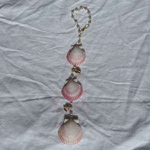 May include: A hanging ornament featuring a circular loop of small white beads connected to a vertical strand. This strand includes three fan-shaped seashells with white and pink gradient coloring, interspersed with small white beads and gold-toned accents.
