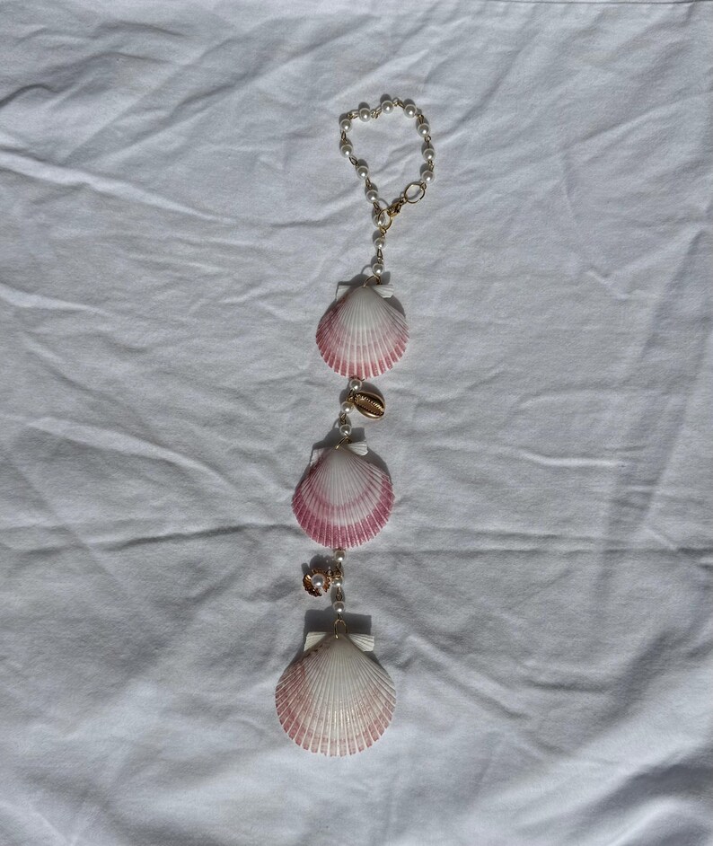 May include: A hanging ornament featuring a circular loop of small white beads connected to a vertical strand. This strand includes three fan-shaped seashells with white and pink gradient coloring, interspersed with small white beads and gold-toned accents.