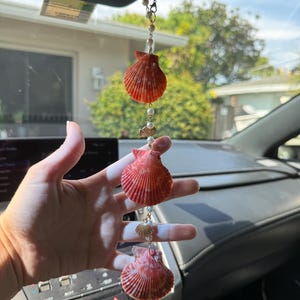 May include: A hanging car ornament featuring three orange-red scallop shells arranged vertically, interspersed with small white faux pearls and tiny gold-toned metal fish and shell charms, all connected by a thin gold-toned metal chain.