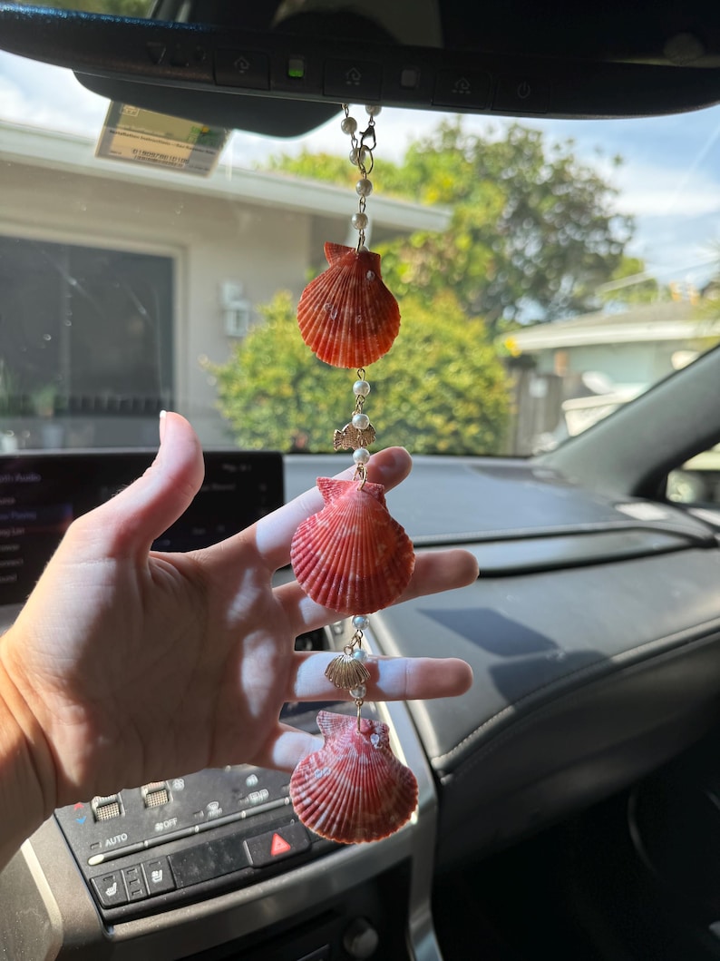 May include: A hanging car ornament featuring three orange-red scallop shells arranged vertically, interspersed with small white faux pearls and tiny gold-toned metal fish and shell charms, all connected by a thin gold-toned metal chain.