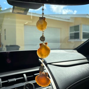 May include: A hanging car ornament featuring three orange scallop shells arranged vertically, interspersed with small white beads and two circular metal charms, suspended from a delicate gold-toned chain attached to a rearview mirror.