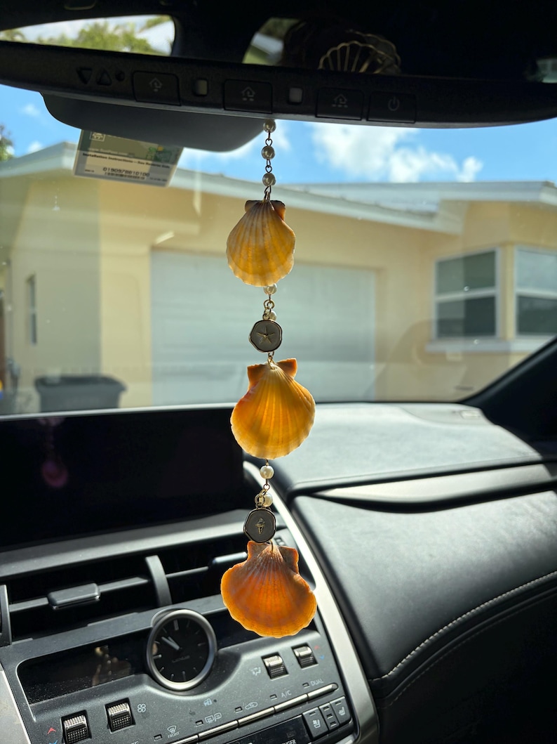 May include: A hanging car ornament featuring three orange scallop shells arranged vertically, interspersed with small white beads and two circular metal charms, suspended from a delicate gold-toned chain attached to a rearview mirror.