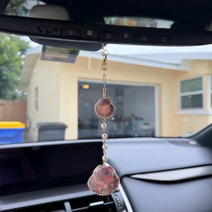 May include: A hanging car ornament featuring two scalloped seashells in shades of brown and tan, connected by a gold-toned metal chain and a series of small white spherical beads, suspended from a rearview mirror.