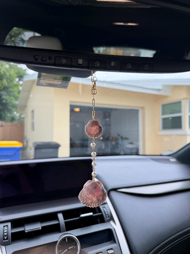 May include: A hanging car ornament featuring two scalloped seashells in shades of brown and tan, connected by a gold-toned metal chain and a series of small white spherical beads, suspended from a rearview mirror.
