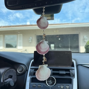 May include: A car rearview mirror hanging ornament featuring three white and reddish-pink scallop shells strung together with small white beads on a gold-toned chain. The shells are arranged vertically, separated by the beads and small gold accents.