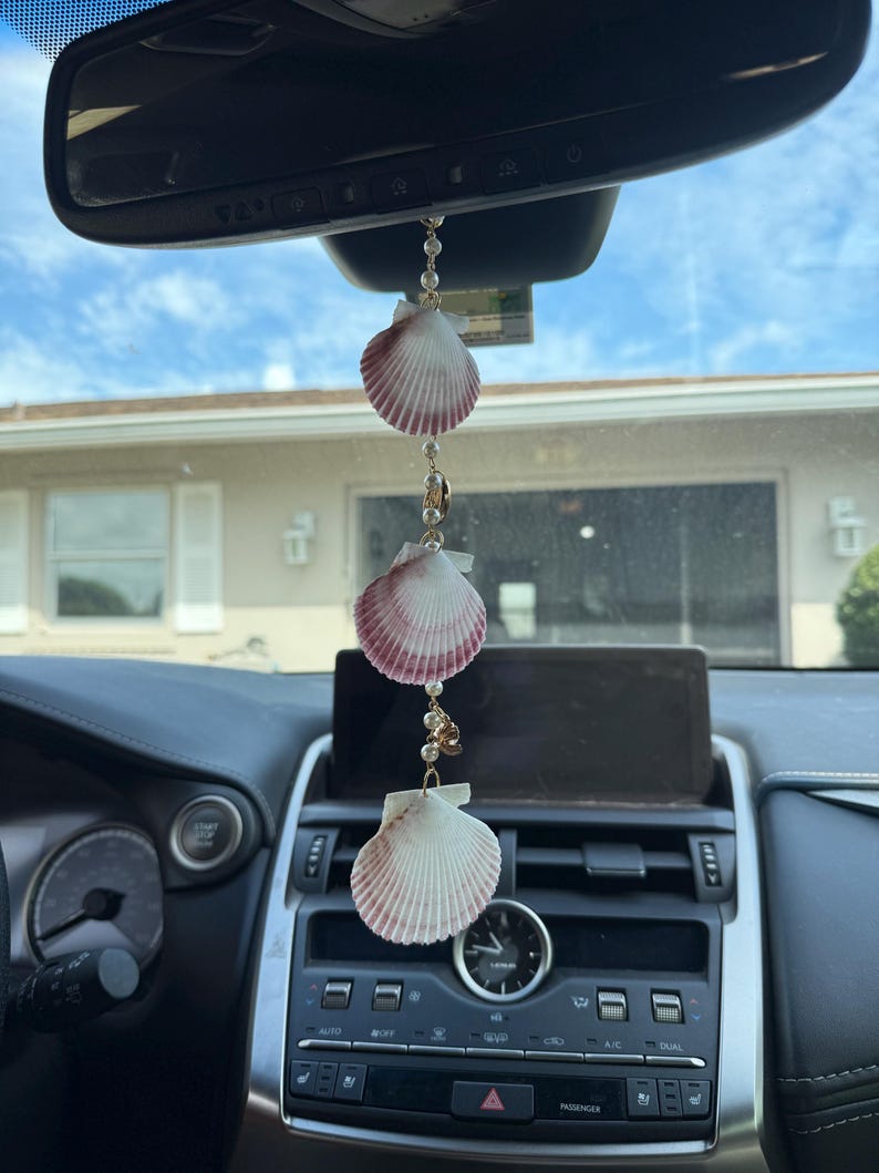 May include: A car rearview mirror hanging ornament featuring three white and reddish-pink scallop shells strung together with small white beads on a gold-toned chain. The shells are arranged vertically, separated by the beads and small gold accents.
