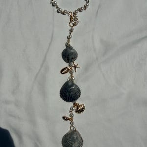 May include: A hanging ornament featuring a circular loop of white faux pearls connected to a vertical chain. The chain is adorned with three large grey ridged seashells, a small gold-toned starfish charm, and two smaller gold-toned shell charms.