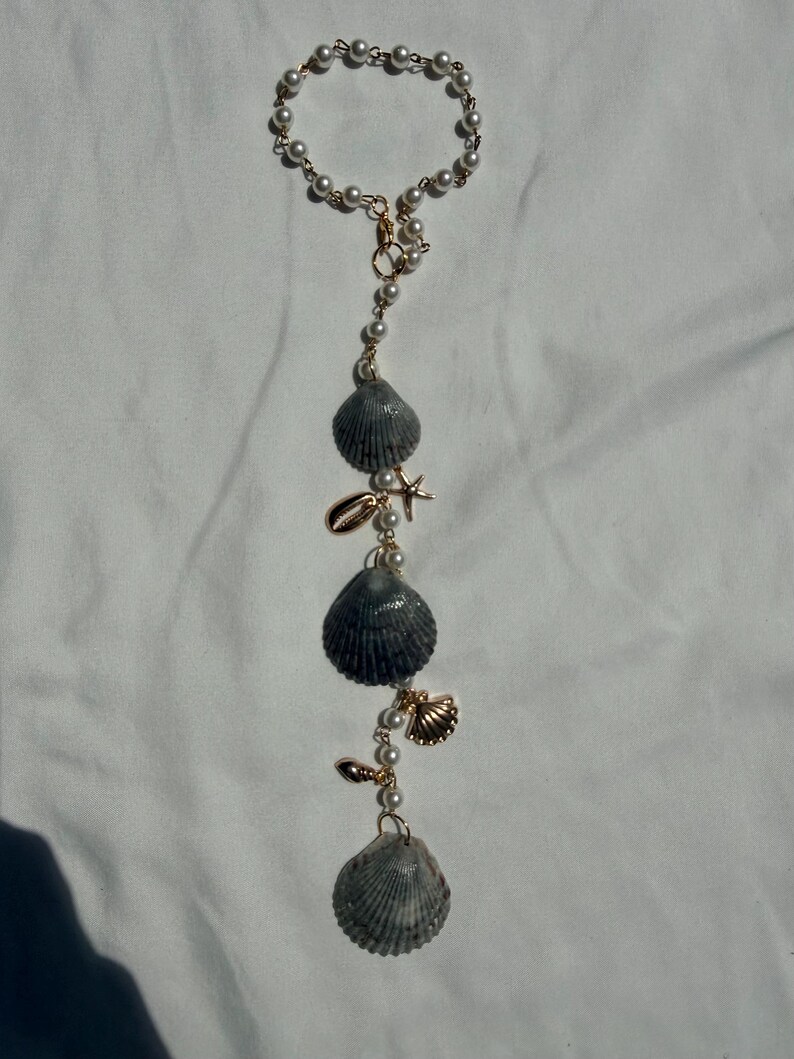 May include: A hanging ornament featuring a circular loop of white faux pearls connected to a vertical chain. The chain is adorned with three large grey ridged seashells, a small gold-toned starfish charm, and two smaller gold-toned shell charms.