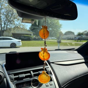 May include: A hanging car ornament featuring three orange and yellow scallop shells arranged vertically, connected by a silver chain with small white beads and metallic charms, suspended from the rearview mirror of a vehicle.