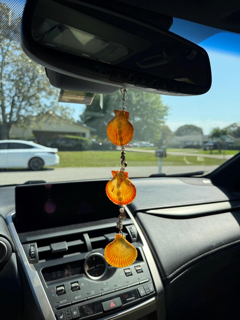 May include: A hanging car ornament featuring three orange and yellow scallop shells arranged vertically, connected by a silver chain with small white beads and metallic charms, suspended from the rearview mirror of a vehicle.