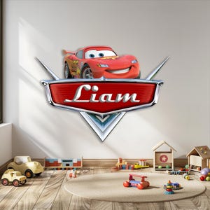 May include: A red Lightning McQueen car decal with the name "Liam" on a red and silver emblem. The decal is above a collection of toy cars and wooden blocks on a light brown rug.