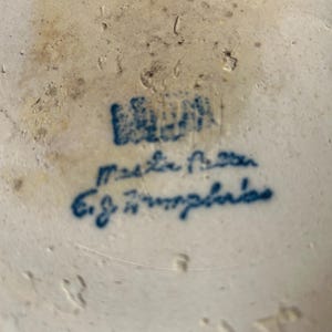 Może przedstawiać: Kremowa ceramiczna podstawa z niebieskim nadrukiem i odręcznym tekstem wykonanym niebieskim tuszem. Powierzchnia wykazuje oznaki starzenia z jasnobrązowymi przebarwieniami i niewielkimi wgłębieniami w strukturze materiału ceramicznego.