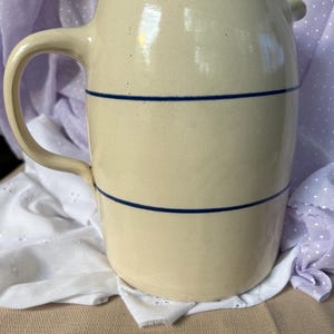 May include: A cream-colored ceramic pitcher with a smooth, matte finish. The vessel features a single curved handle on the left side and two distinct, thin horizontal navy blue stripes encircling the body of the pitcher, providing a simple contrast.