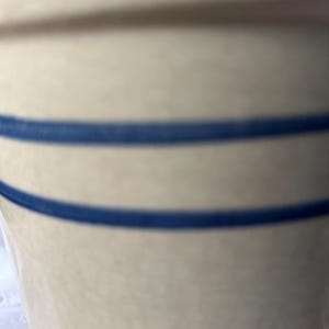 Puede incluir: Una vasija de gres cilíndrica con un acabado liso de color crema blanquecino. El exterior presenta dos franjas azules horizontales y paralelas distintas que rodean el cuerpo del recipiente, creando un contraste con la base neutra.