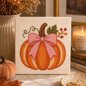 May include: A square cross-stitch embroidery on white fabric featuring an orange pumpkin with a brown stem, a green leaf, and a light pink bow tied around the center. The design includes a small green vine and a cluster of orange and red berries.