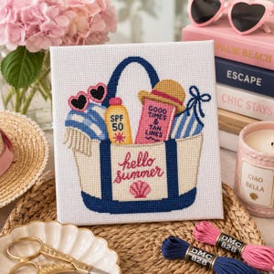 May include: A square cross-stitch embroidery on white fabric featuring a cream-coloured tote bag with navy blue handles. The bag contains pink heart-shaped sunglasses, a yellow SPF 50 bottle, a pink book titled Good Times & Tan Lines, and a straw hat.