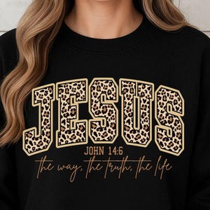 May include: Black sweatshirt featuring the word "JESUS" in leopard print lettering. The text "JOHN 14:6" and "the way, the truth, the life" are printed below the word "JESUS" in a gold colour.