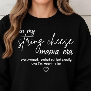 Könnte beinhalten: Schwarzes Sweatshirt mit weißem Text: "in my string cheese mama era". Der Text darunter lautet "overwhelmed, touched out but exactly who I'm meant to be" mit einem kleinen Herzen. Das Sweatshirt hat einen lässigen Stil.