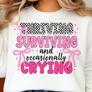 May include: White sweatshirt with the text "THRIVING, SURVIVING, and occasionally CRYING" in a mix of fonts and colors. The word "THRIVING" is in a black and white checkered pattern. The word "SURVIVING" is in pink leopard print.