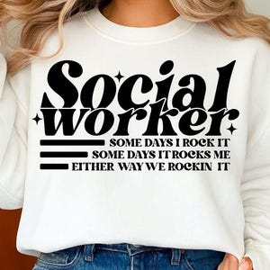May include: White sweatshirt with the words "Social Worker" in a bold, black font. Below the text are the phrases "SOME DAYS I ROCK IT", "SOME DAYS IT ROCKS ME", and "EITHER WAY WE ROCKIN IT". The sweatshirt is paired with blue jeans.