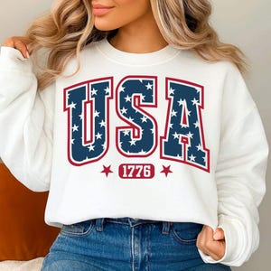 Pode incluir: Camisola branca com a palavra "USA" em letras azul marinho preenchidas com estrelas brancas, contornadas a vermelho. Abaixo da palavra "USA" está o número "1776" num retângulo vermelho, ladeado por estrelas vermelhas.