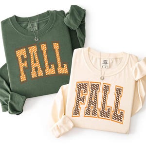 Könnte beinhalten: Ein langärmeliges Sweatshirt in Dunkelolivgrün und ein langärmeliges Sweatshirt in Creme, beide mit dem Wort FALL in großen Blockbuchstaben. Das grüne Oberteil hat orangefarbene Buchstaben mit weißen Punkten, das cremefarbene Oberteil hat braune Buchstaben mit weißen Punkten.
