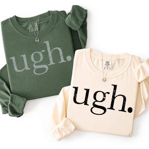 May include: A dark olive green long-sleeve sweatshirt and a cream long-sleeve sweatshirt, both featuring the word "ugh." printed in a distressed, weathered font across the chest. Each garment has a crew neckline and a visible interior brand label.