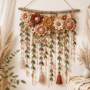 May include: A wall hanging featuring a horizontal wooden branch adorned with various crocheted flowers in shades of rust, cream, peach, and sage green. Multiple strands of crocheted leaves, small flowers, and wooden beads hang down with tassels.