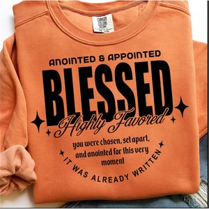 May include: An orange sweatshirt with the words "ANOINTED & APPOINTED BLESSED Highly Favored" in black. The text "you were chosen, set apart, and anointed for this very moment. It was already written" is also printed on the sweatshirt.