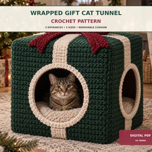 Op de afbeelding: Een donkergroene gehaakte kubusvormige kattentunnel in de vorm van een ingepakt cadeau, met een crèmekleurig lintpatroon, een rode strik bovenop en twee ronde ingangen. Een cyperse kat zit erin. Tekst: WRAPPED GIFT CAT TUNNEL CROCHET PATTERN.
