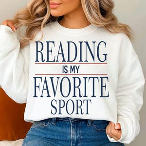May include: White sweatshirt with the text "READING IS MY FAVORITE SPORT" in navy blue, with a red line separating the words. The sweatshirt is paired with blue jeans.