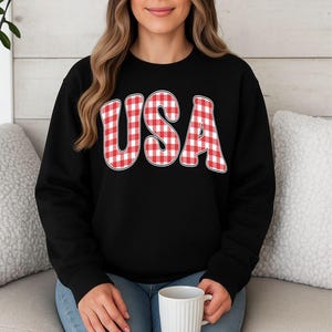 Peut inclure: Un sweat-shirt noir à col rond arborant le texte USA dans un motif vichy rouge et blanc. Les lettres présentent une police arrondie et audacieuse avec un fin contour blanc, centrées sur la poitrine du vêtement à manches longues.