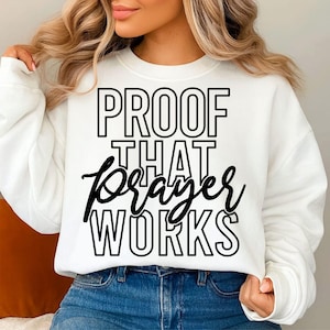 May include: White sweatshirt with the text "PROOF THAT prayer WORKS" in black. The text is in a bold, outlined font, with the word "prayer" in a cursive script. The sweatshirt is a casual, comfortable style.