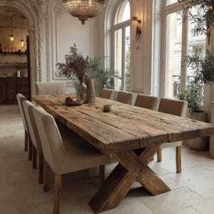 Puede incluir: Una mesa de comedor rústica de madera con base de patas cruzadas, rodeada de sillas tapizadas. La mesa es de madera recuperada y tiene un acabado natural y envejecido. Una lámpara de araña cuelga sobre la mesa. La mesa mide unos 300 cm de largo.