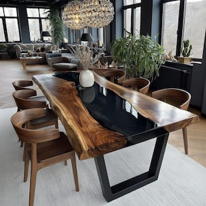 Op de afbeelding: Een grote eettafel met een natuurlijke rand en een zwarte epoxyhars inleg. De tafel heeft een natuurlijke houtnerf en staat op een zwart metalen onderstel. Er staan meerdere houten stoelen om de tafel.
