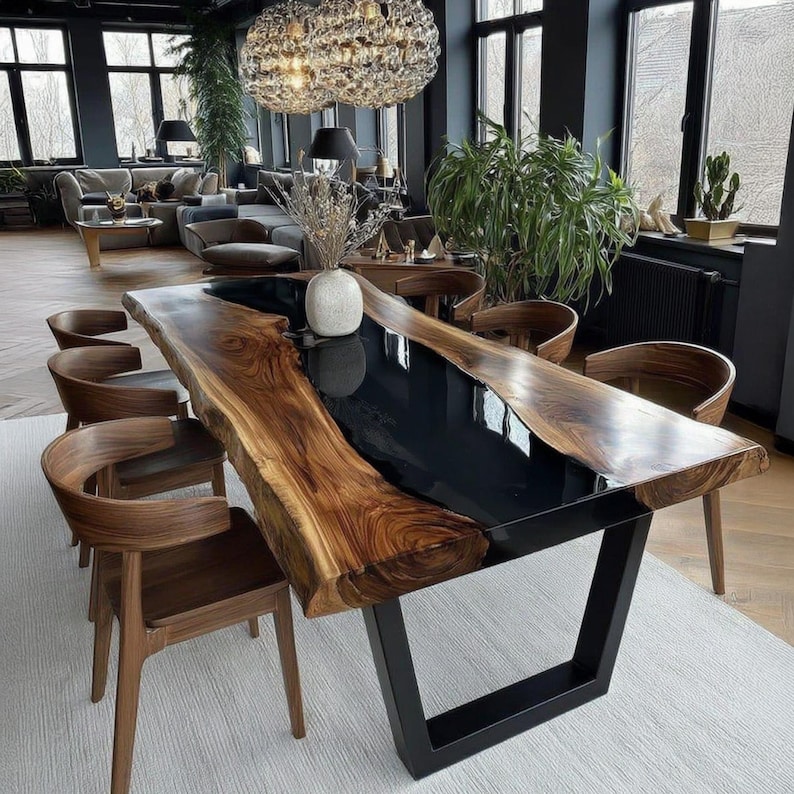 Op de afbeelding: Een grote eettafel met een natuurlijke rand en een zwarte epoxyhars inleg. De tafel heeft een natuurlijke houtnerf en staat op een zwart metalen onderstel. Er staan meerdere houten stoelen om de tafel.